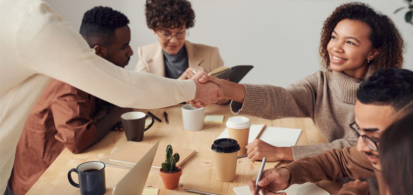 A person sitting at the right-hand side of the table shakes hands with someone who is standing across from them.