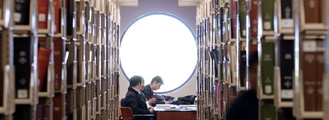 A view in between library bookshelves that leads to three people studying at the end of room at a table by a window.