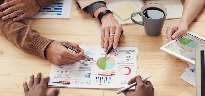 Economics. A close-up of a paper on a table with various graphs and charts. A close-up of a paper on a table with various graphs and charts. People’s hands are pointing to different charts while holding pens.