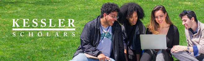 A group of people sitting on grass outdoors looking at a laptop.