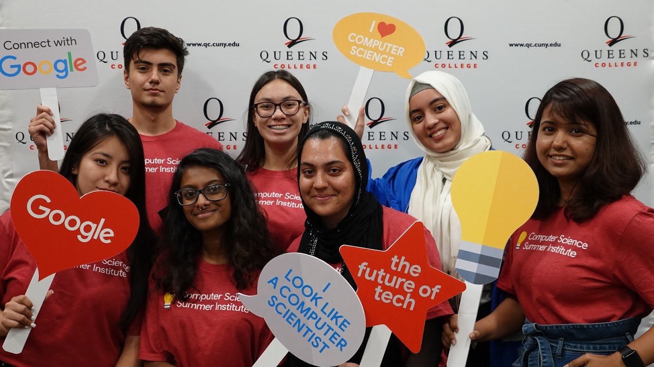 Group photo of participants holding various photobooth prop signs. Group photo of participants holding various photobooth prop signs and posing in front of a Queens College background.