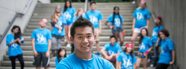 Macaulay Honors College. In the foreground, a person stands close and smiles for the camera. In the background, the group of students sitting or standing by the steps are out of focus. In the foreground, a person stands close and smiles for the camera. In the background, the group of students sitting or standing by the steps are out of focus.