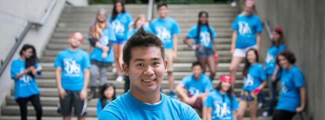 In the foreground, a person stands close and smiles for the camera. In the background, the group of students sitting or standing by the steps are out of focus.