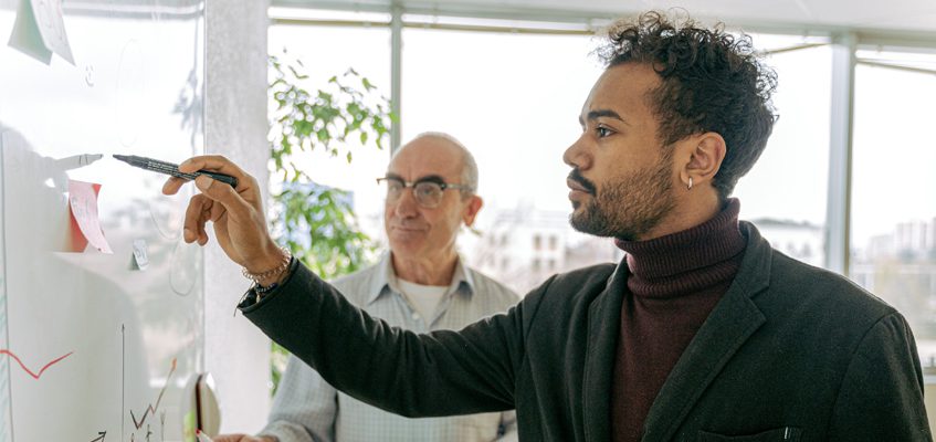 Two people standing in front of a white board. One person is writing, and one person is observing.