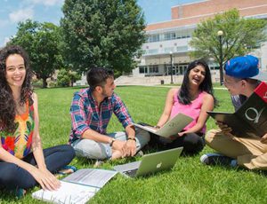 A group of people sitting on the grass at Queens College Campus. They have laptops in front of them and are having a conversation. A group of people sitting on the grass at Queens College Campus. They have laptops in front of them and are having a conversation.