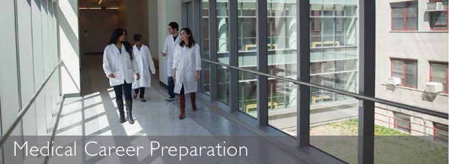 Four people in lab coats walk through a hallway with windows.