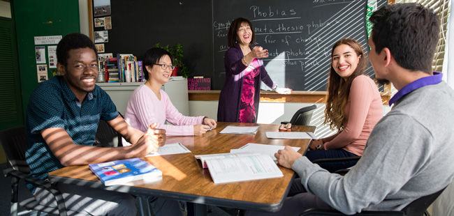 A teacher standing by the blackboard gesturing towards her students with her hand. Four students are sitting down at the table in front of the blackboard with their textbooks opened.