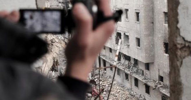 A close-up of a person’s hand holding a video camera. They are recording a partially demolished building.
