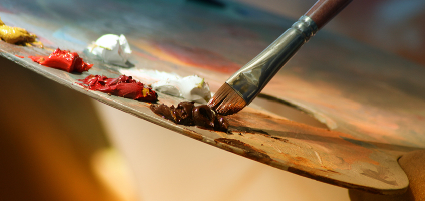 A close-up of a brush being dipped into an artist’s paint palette.