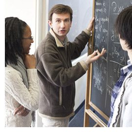 A person speaking while pointing to a chalkboard with two people observing him.