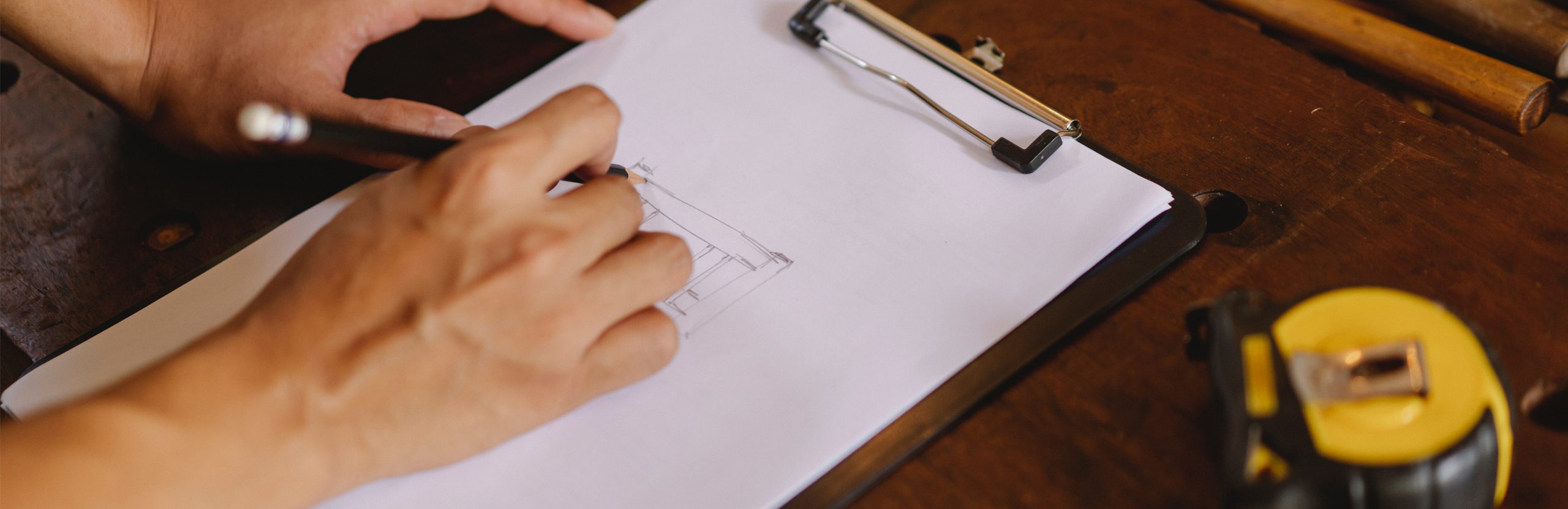 A close-up of a person’s hand drawing a diagram on a clipboard. There is a tape measure on the right-hand side of the table.