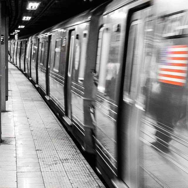 Black and White image of a NYC train in motion. The American flag on the side of the train is in color.