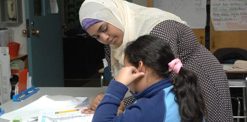Hasina Islam stands next to a child and helps them with their homework.