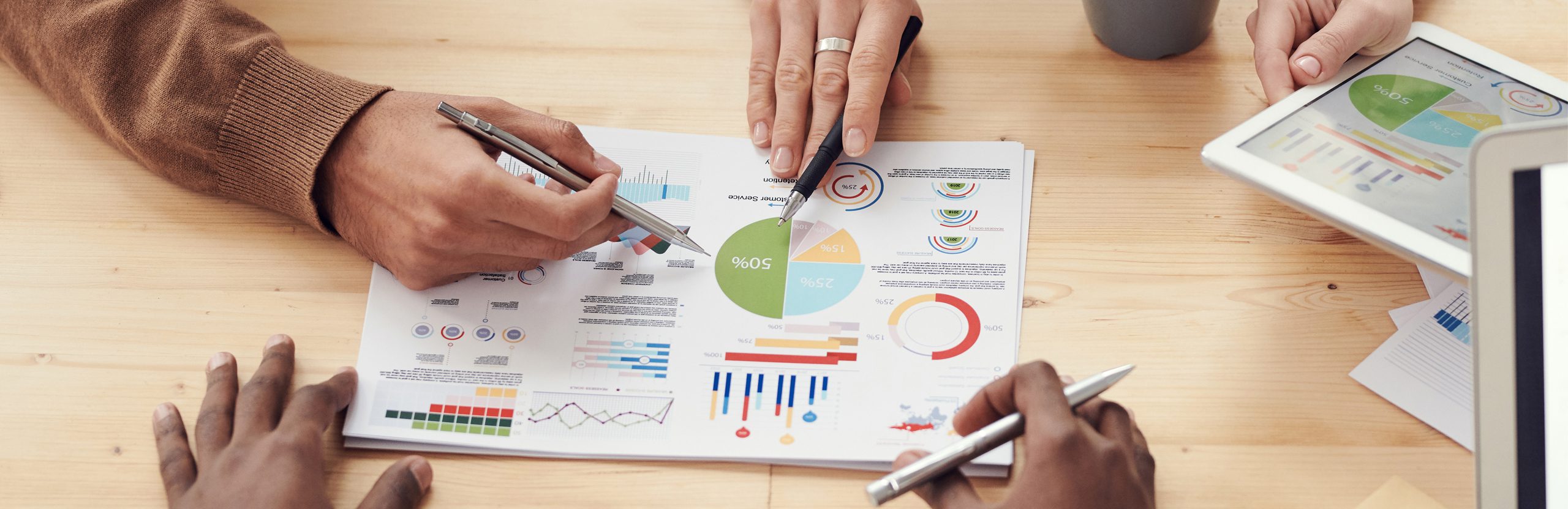 A close-up shot of a table with a paper that contains graphs and charts. Various hands from all sides of the image hold pens as they point to different parts of the paper.