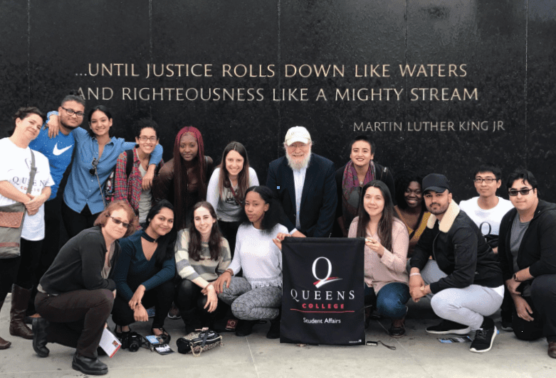 A group photo of the “Footsteps of Dr. King” trip. The people are posing in two rows. The first row is kneeling. Some people in the second row are crouched over while some are standing. In the background a quote is written on the wall. It reads “…Until justice rolls down like waters and righteousness like a mighty stream” – Martin Luther King Jr.