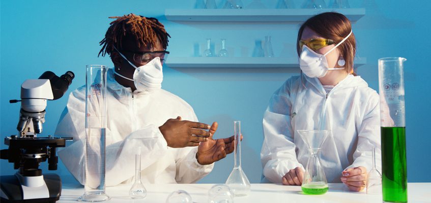 Two people wearing face masks are sitting at a lab table and talking.