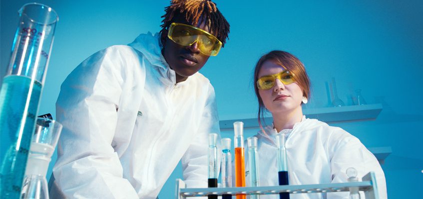 Two people in a lab wearing yellow safety goggles and protective gear look towards the camera.