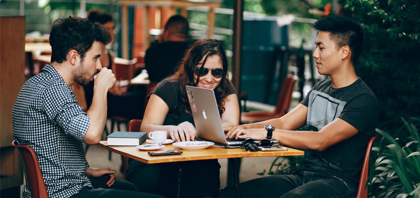 Three people sitting at a table in a common area drinking coffee and working on a laptop.