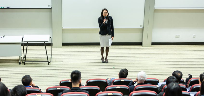 A person standing in front of a lecture hall while holding a microphone.