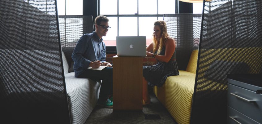 Two people sitting on opposite sides share a laptop on the table in the center.