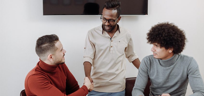 Two people sitting down, the person on the left is shaking hands with a person standing between them.