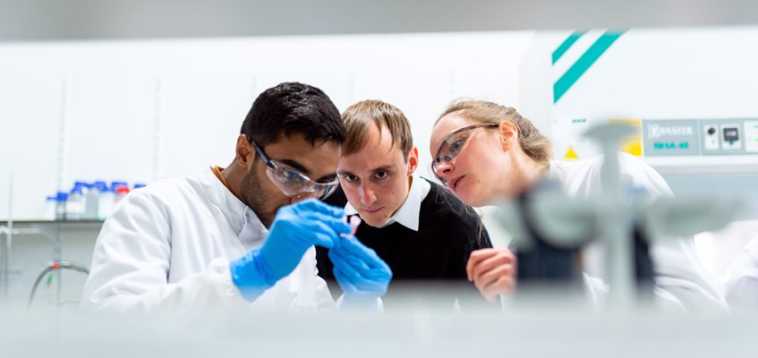 Three people standing in a lab together observing a vial.