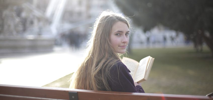 A person sitting on a bench with their back facing us. They have their head turned looking towards the camera while holding a book.