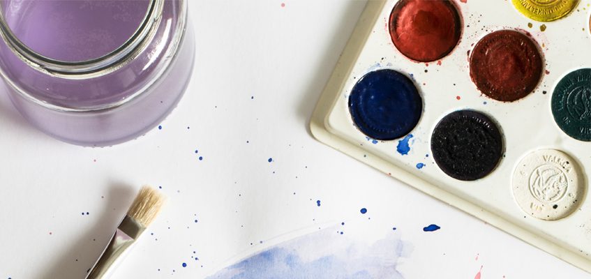Watercolor palette box, paint brush, and glass paint water cup rest on a slightly paint splattered white surface.