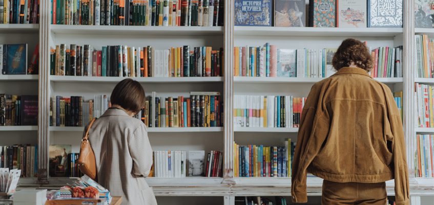 Two people with their backs facing the camera look at a bookshelf.