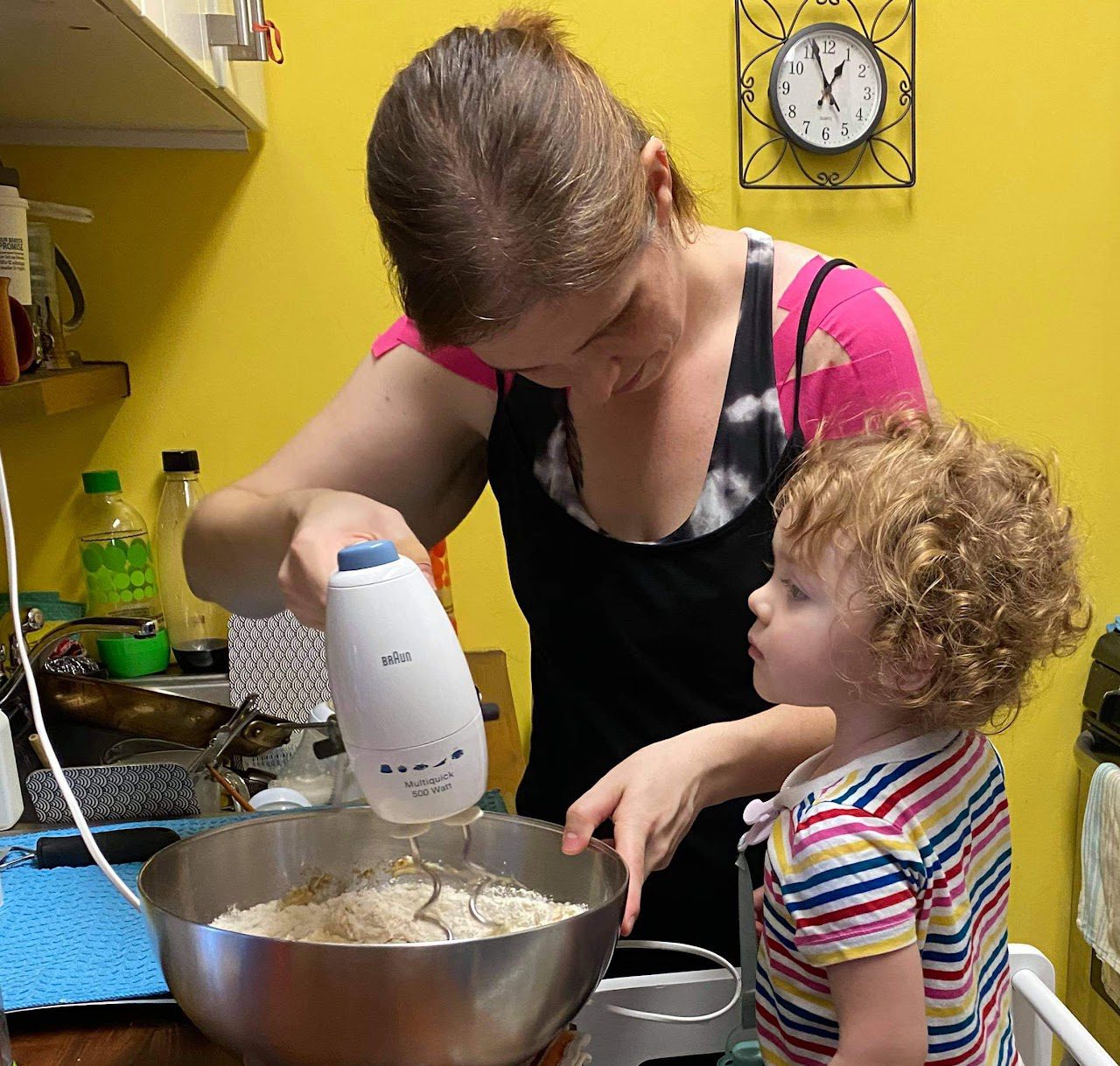 Corinna Singleman using an electric hand mixer on dough in a large metal bowl. A child pays attention attentively on the side.