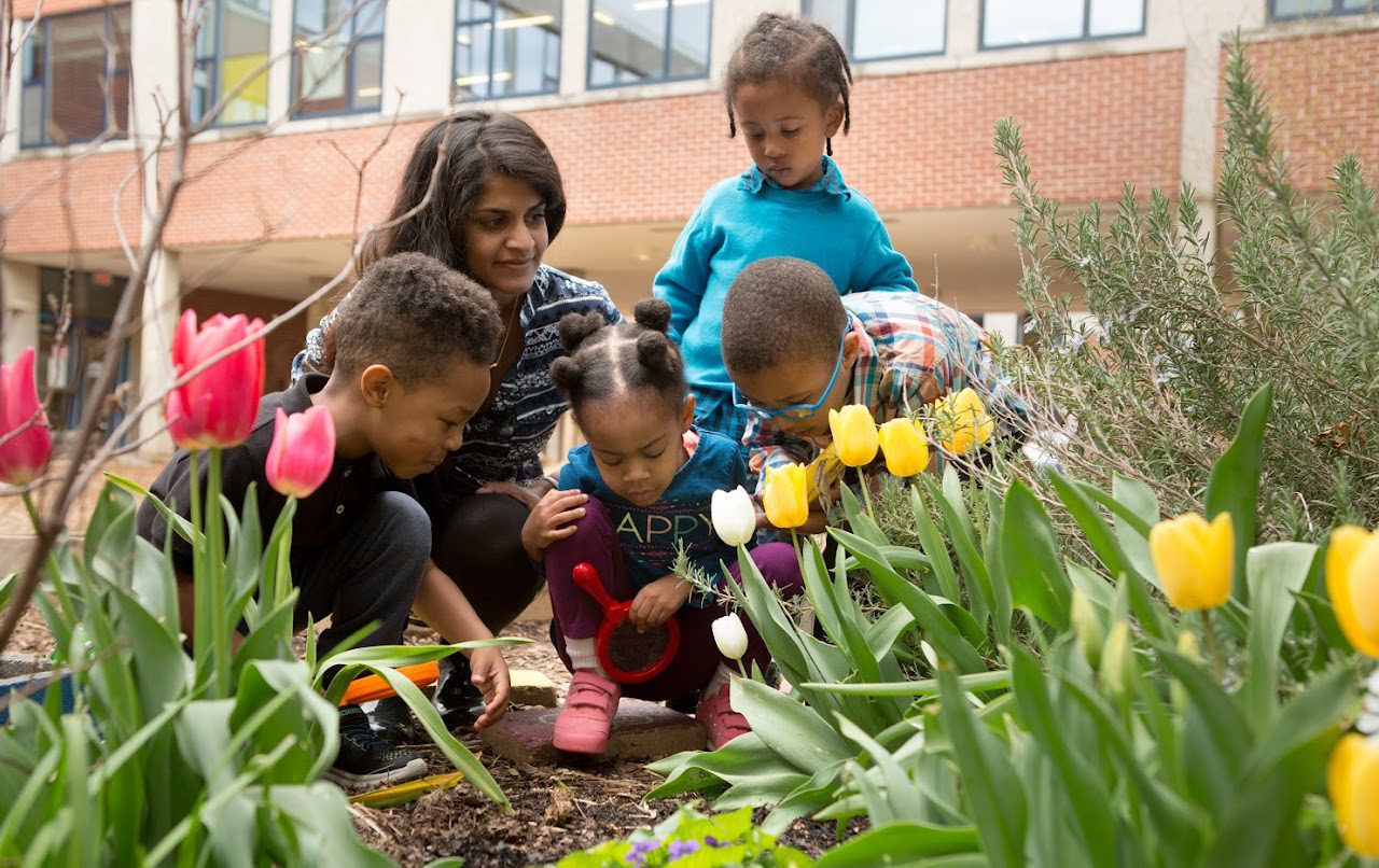 A teacher with four students explore a garden together. They are all focused on something in the soil.