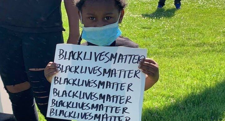 A young child holding up a poster that reads “Black Lives Matter” 6 times.