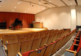 A view of an empty Recital Room. It features the seating area and a view of the stage which contains a baby grand piano.