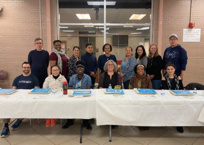 Two rows of people. The first row is sitting at a table with a white tablecloth and books. The second row is standing behind them.