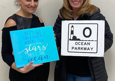 Two people holding signs. The first sign reads: Let’s go somewhere where the stars meet the ocean. The second sign is an Ocean parkway Road Sign.