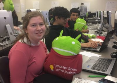 Students sitting by computers holding the Android stuffed animal mascot.