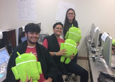 Three students sitting near their computers holding the Android stuffed animal mascot.