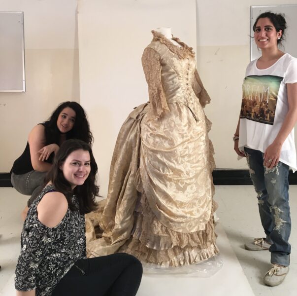 Three students standing around a late 18th century champagne gown of brocade silk and lace.