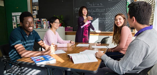 Center for Academic and Communication Skills. A teacher standing by the blackboard gesturing towards her students with her hand. Four students are sitting down at the table in front of the blackboard with their textbooks opened.