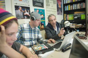 Photo of students learning podcasting from their professor. The image shows two students in the foreground with headphones on and professor Douglas Rushkoff pointing at a computer screen