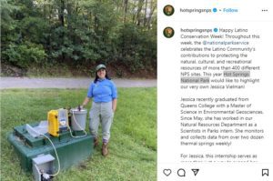 A woman with dark hair wearing a blue shirt, light green pants and a green baseball cap stands in a grassy field next to a green cement box with a grey bucket and yellow case on top