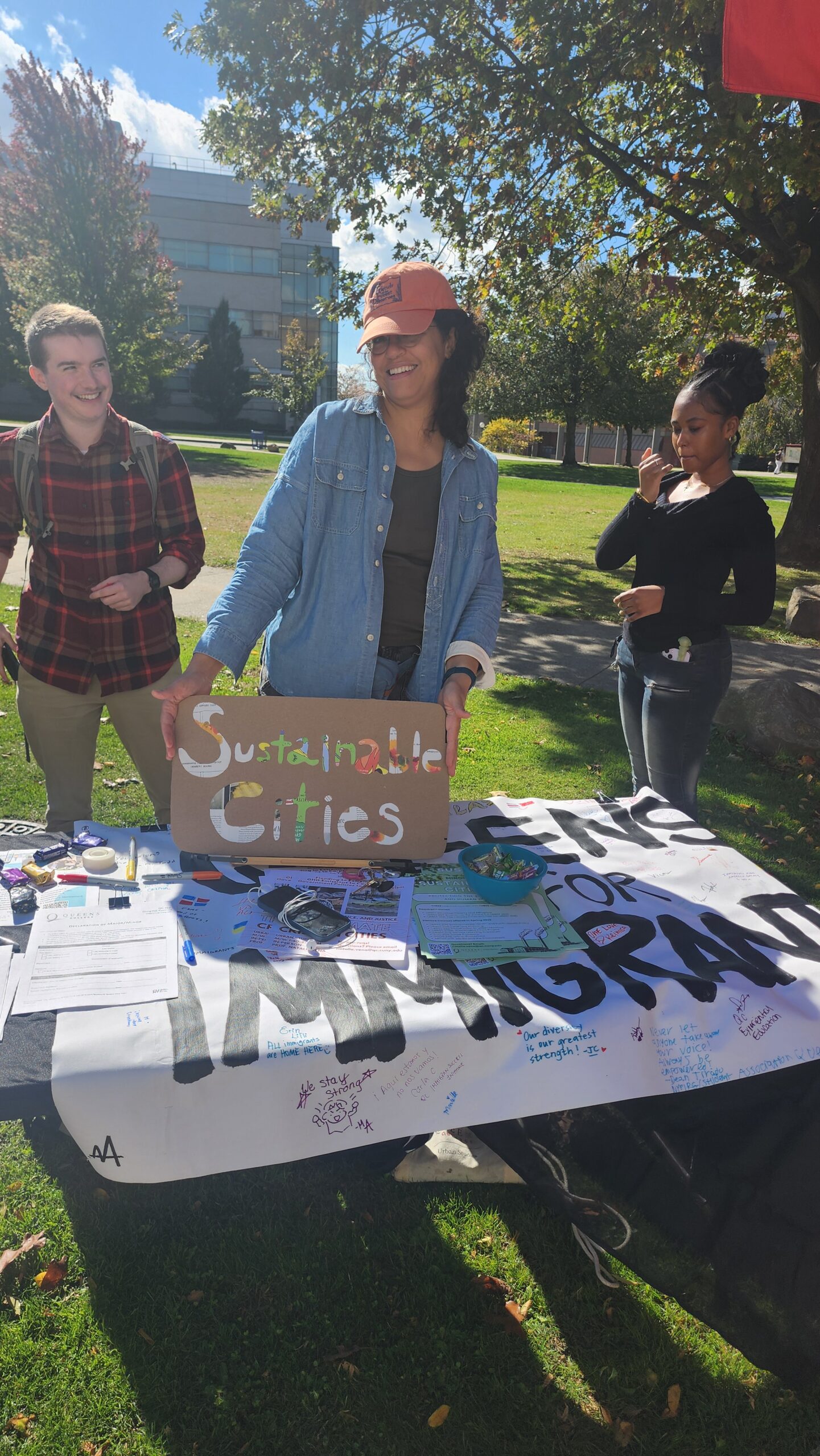 Three people standing behind a table. The person in the middle holds up a sign reading “Sustainable Cities”.