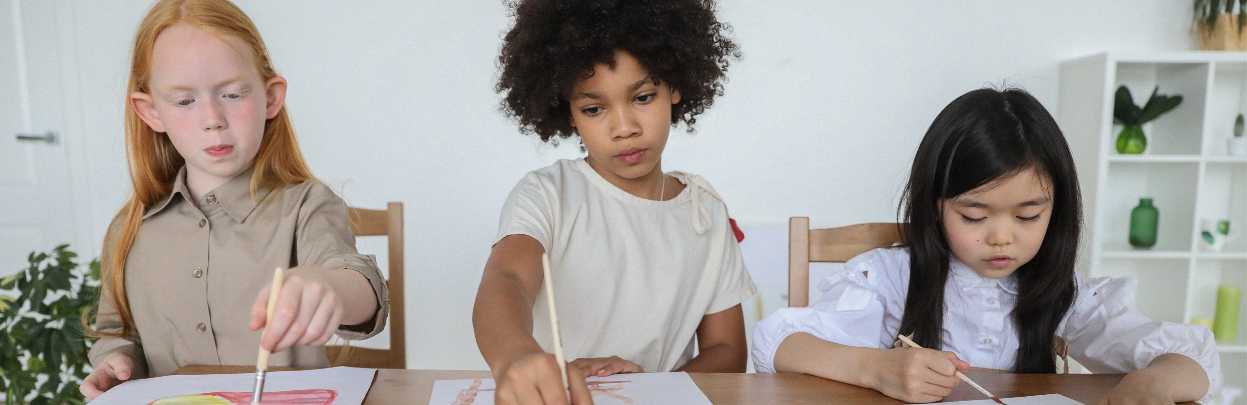 Three children are sitting at a desk and painting together.