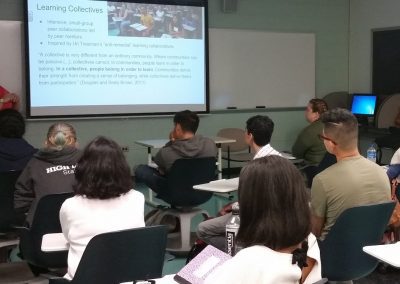 A group of people sitting in a classroom focused on the lecturer and the slides on the projector screen.