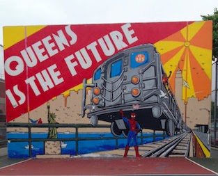 A mural on a handball court at IS 145 of Spiderman holding up a train. A mural on a handball court at IS 145 of Spiderman holding up a train. The text reads “Queens Is the Future”.
