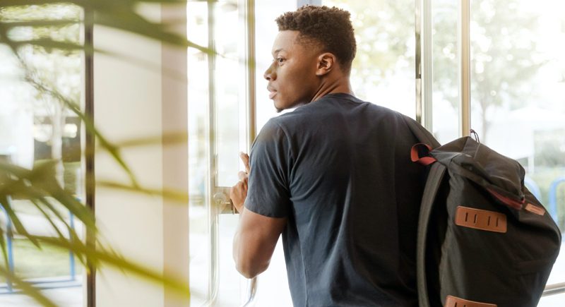 A person carrying a backpack while holding onto the handle of a glass door.