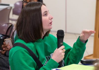 A person in a green sweater holding a microphone and speaking.