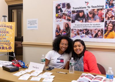 Two people sitting side-by-side behind a table pose for a photo. Behind them is a poster for transitions to teaching.