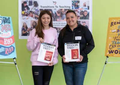 Two people standing side-by-side holding the book “Wisdom Tales”. There is a poster on the left-hand side of the room and on the right-hand side of the room.