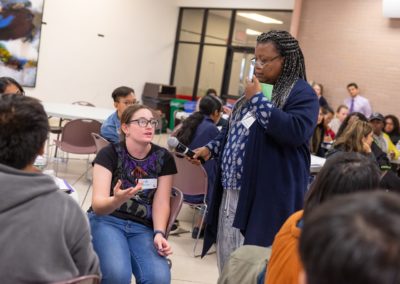 A person standing and holding the microphone lowered for a person sitting and speaking.